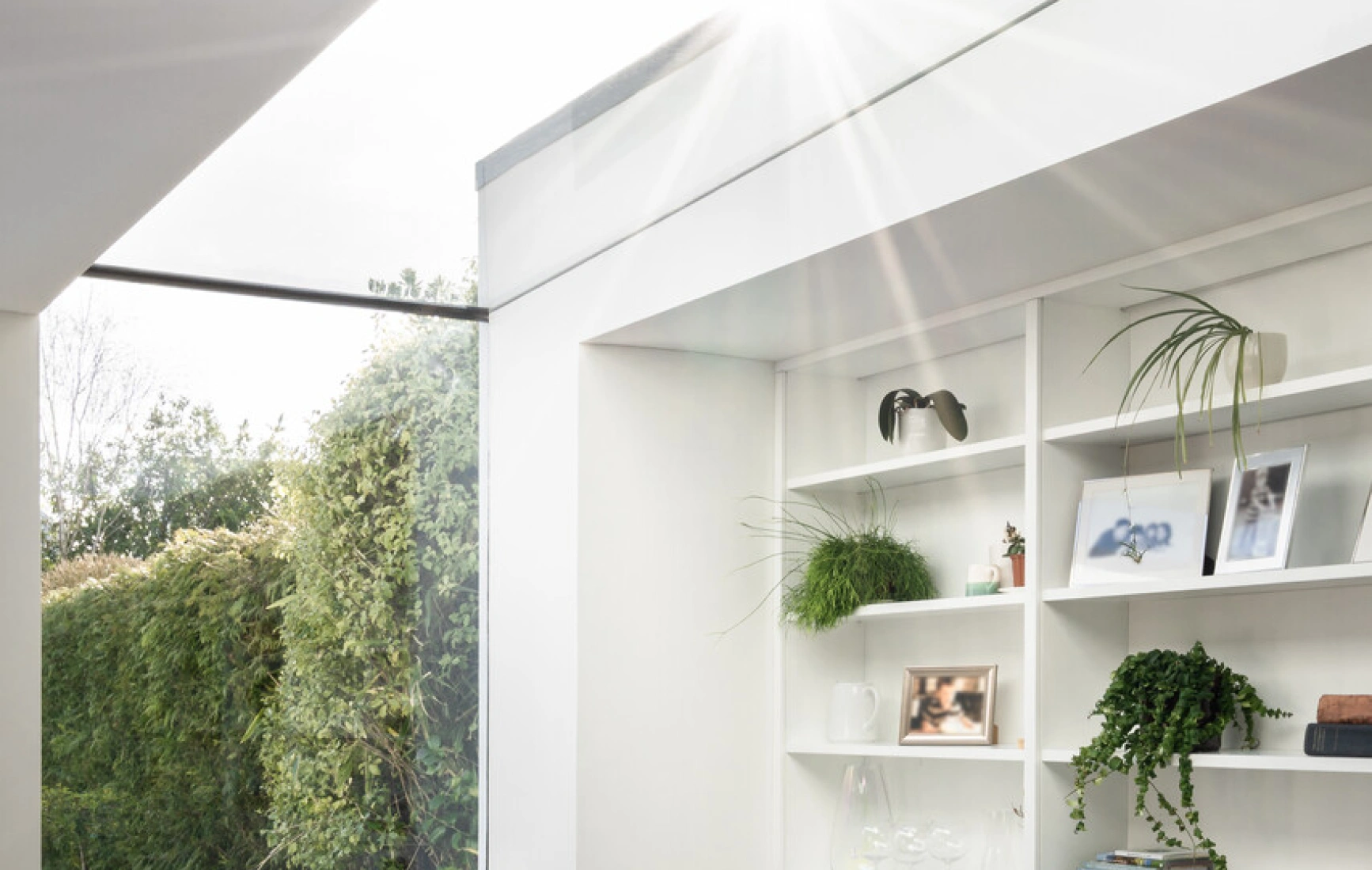Heller Wohnraum mit einem Fenster, das Licht ins Zimmer lässt. Eine bequeme Sessel steht neben einem Bücherregal mit Pflanzen. Draußen ist ein grüner Garten sichtbar.