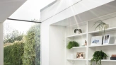 Un coin lumineux d'une pièce moderne avec une chaise confortable, des étagères blanches contenant des plantes et des objets décoratifs. De grandes fenêtres laissent entrer la lumière naturelle, offrant une vue intérieure-extérieur sur un jardin verdoyant.