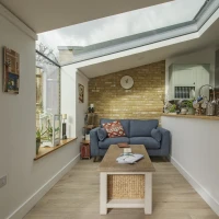 A cozy living space featuring a blue couch, a wooden coffee table with a woven basket, and a brick accent wall. A large skylight allows natural light to fill the room, while plants and decorative items add warmth.