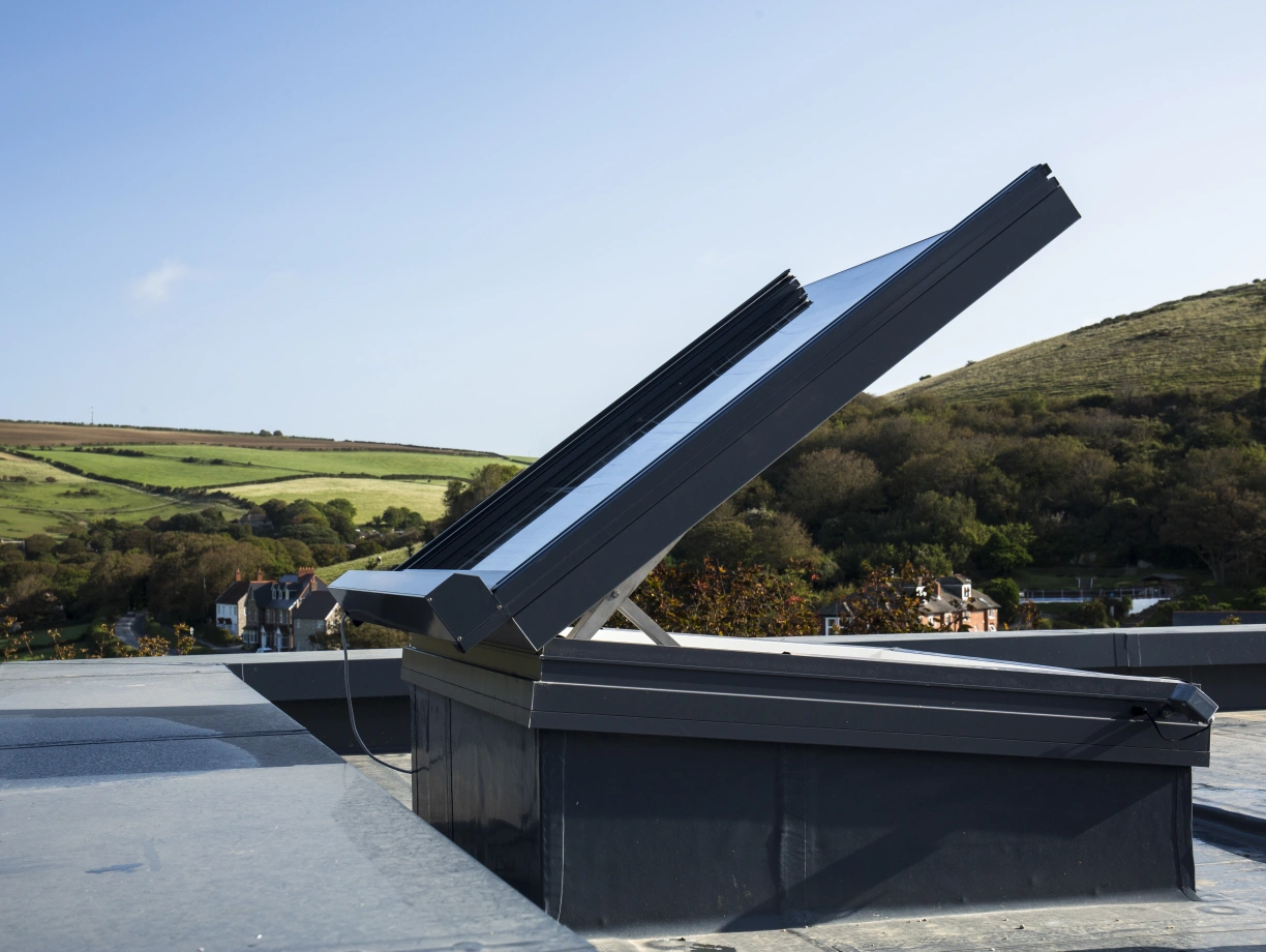 A solar panel is installed on a rooftop, angled to capture sunlight, with a scenic view of hills and greenery in the background. The sky is clear and blue.