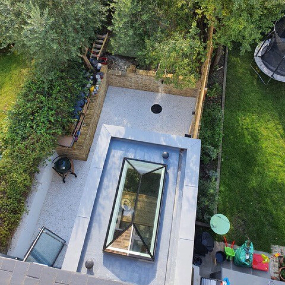 Aerial view of a backyard featuring a patio with a circular hole, surrounded by greenery and trees. There is a small playground area with colorful equipment and a trampoline visible. A glass skylight is seen on the roof in the foreground.