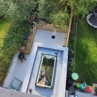 Vista aérea de un jardín con áreas de césped y piedras. Hay un pequeño círculo en el suelo, un patio de grava y un tragaluz en el techo. Al fondo, se ven árboles y un área de juegos.