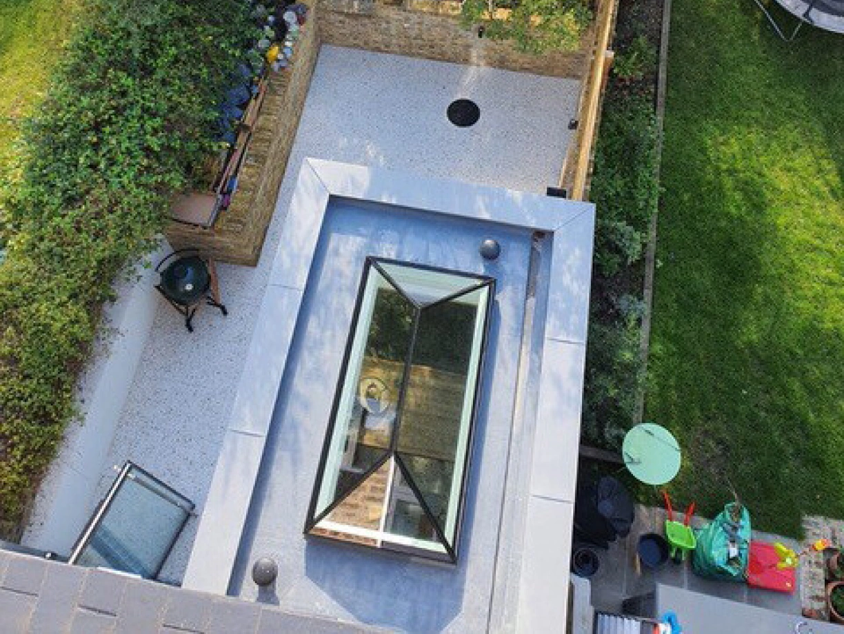 Aerial view of a backyard featuring a patio with a circular hole, surrounded by greenery and trees. There is a small playground area with colorful equipment and a trampoline visible. A glass skylight is seen on the roof in the foreground.