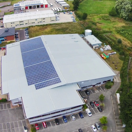 Aerial view of a large commercial building with solar panels on the roof. Surrounding the building are parked cars and a green landscape featuring trees and fields.
