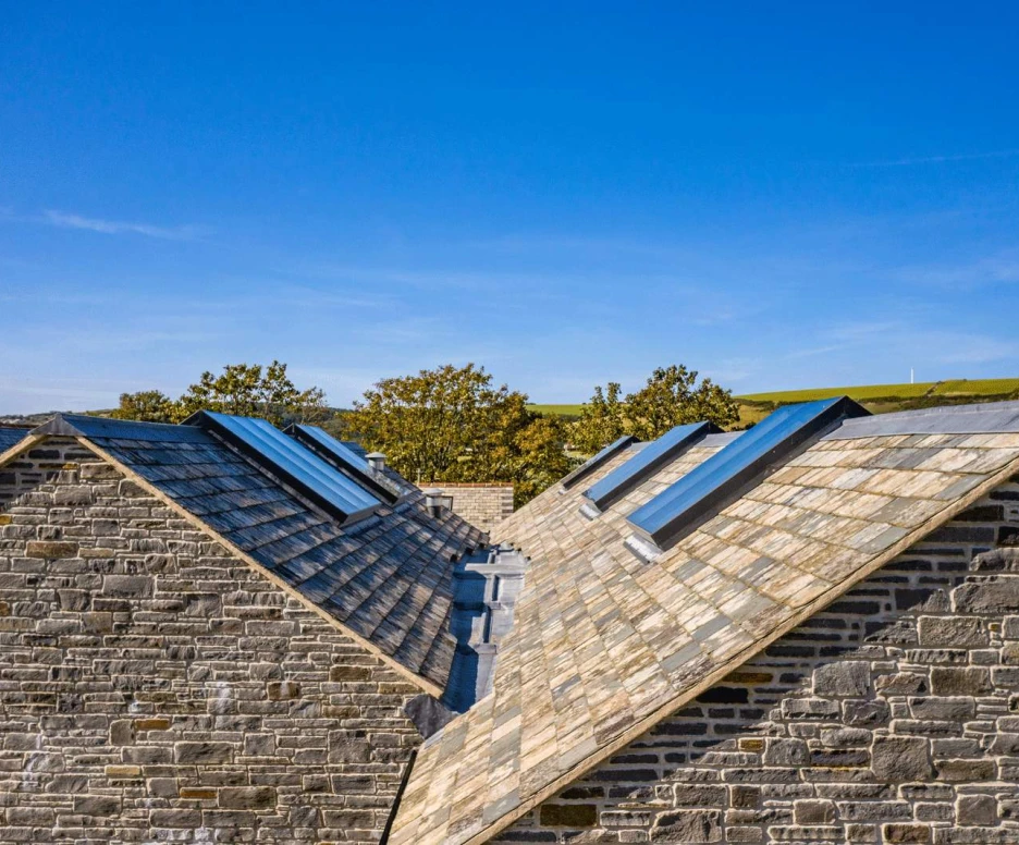 Two sloped rooftops covered in slate tiles, with skylights set into the slopes. Lush green hills are visible in the background under a clear blue sky.