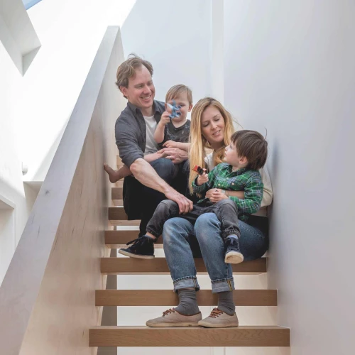 Familia sentada en una escalera de madera. Dos adultos y dos niños pequeños juegan y se miran con alegría. La luz natural entra por una ventana en el techo.