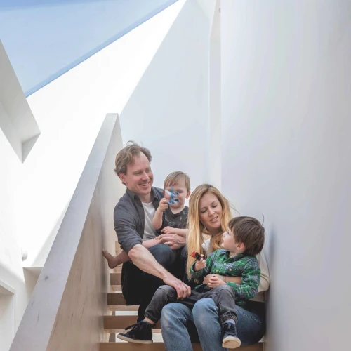 Una familia de cuatro personas sentada en escaleras de madera. Un hombre y una mujer sonríen mientras sostienen a dos niños pequeños. La decoración es moderna y luminosa, con luz natural que entra por una ventana.