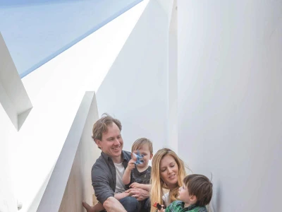 A family of four sits on a wooden staircase. The father holds a young child, while the mother sits with another child on her lap. All are smiling and engaged, with natural light coming from above.