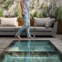 A person walks barefoot over a glass floor section in a modern outdoor space, surrounded by stylish seating and greenery.