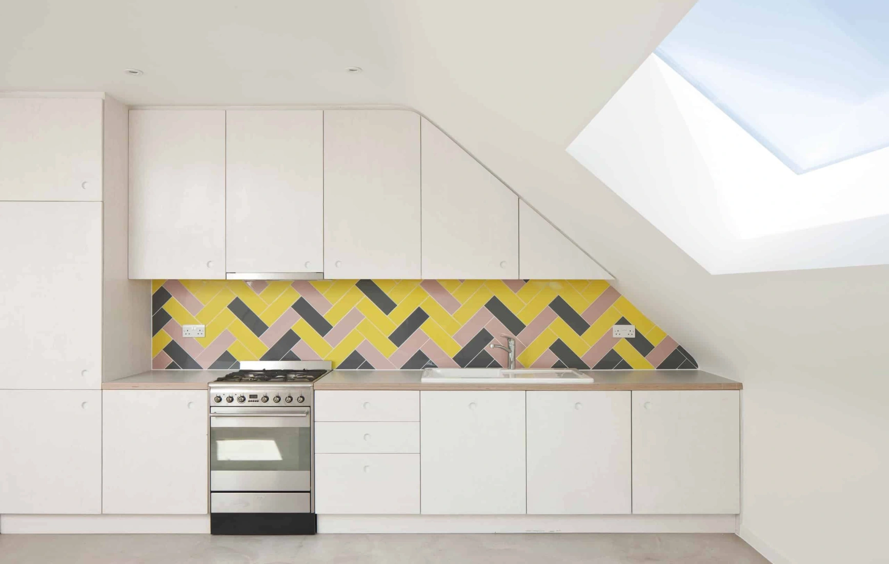 Cocina moderna con armarios blancos, una pared de azulejos en tonos amarillo, rosa y negro, y una ventana en el techo que permite la entrada de luz natural.