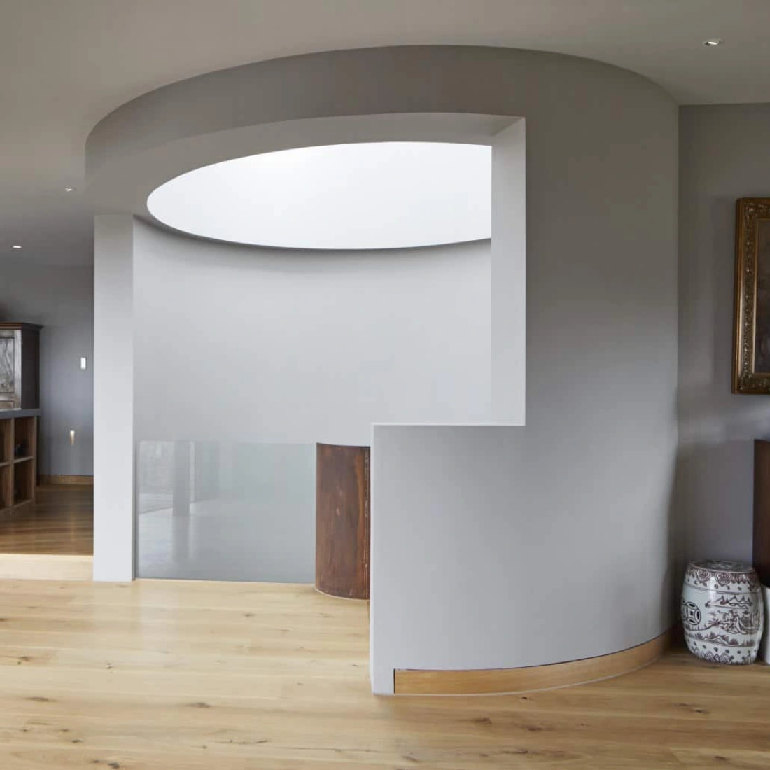 Modern interior space featuring circular architectural design elements, light wooden flooring, and natural light streaming through large glass doors. A dining area with colorful chairs is visible in the background, alongside a wooden cabinet.