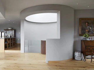 Interior de una casa moderna con paredes grises y suelo de madera. Se observa un espacio abierto que conecta con una cocina y comedor. Hay columnas de madera, un mueble de madera con estantes y una silla de estilo clásico en el fondo. La luz natural entra a través de grandes ventanales.