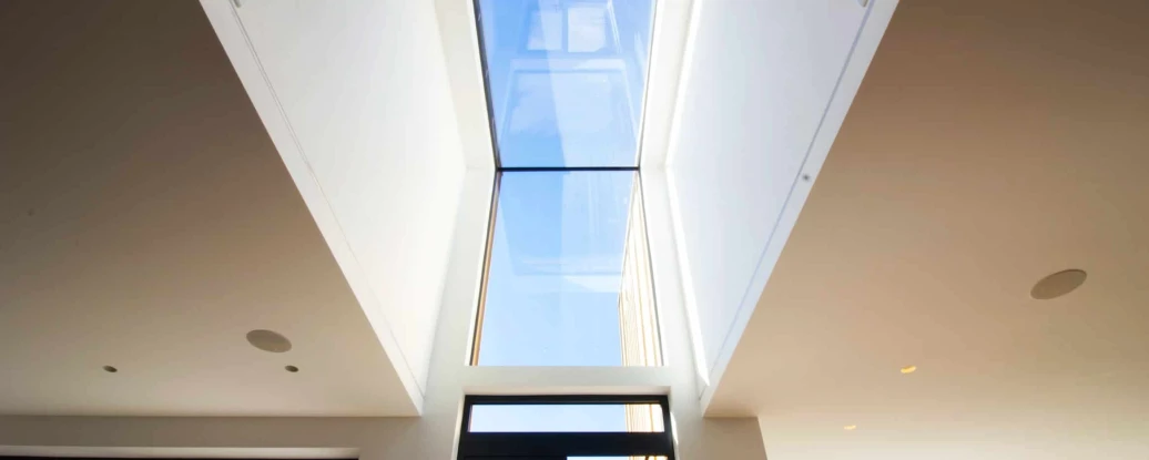 A modern skylight creating a rectangular opening in a white ceiling, allowing natural light to flood in. The view outside shows clear blue skies and greenery. Large windows frame the scene below, enhancing the airy feel of the space.