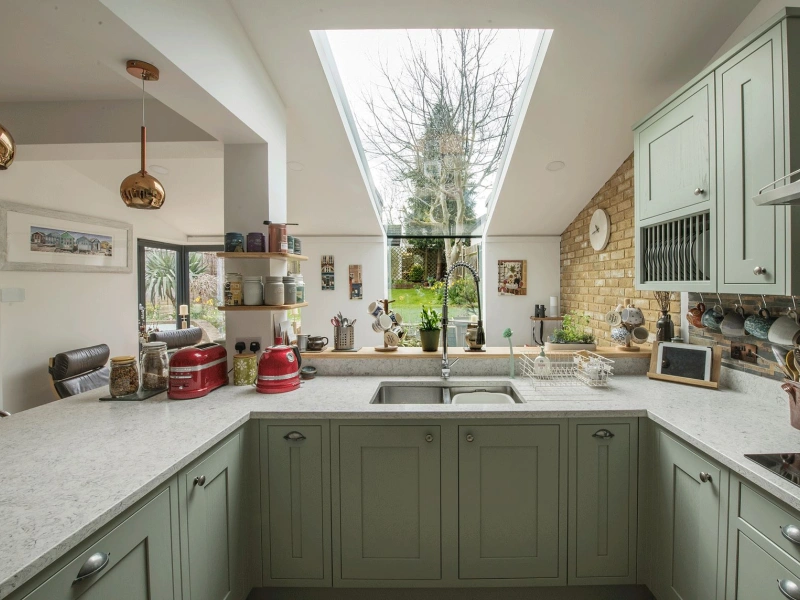 Cuisine moderne avec des armoires vertes, un grand plan de travail en marbre et une fenêtre de toit laissant entrer une belle lumière naturelle. À l’extérieur, le jardin est visible, agrémenté de plantes et de décorations murales en arrière-plan.