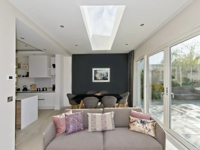 Un salon moderne avec un canapé beige orné de coussins colorés. À l’arrière, une salle à manger avec une table noire et des chaises grises. De grandes fenêtres et un plafond vitré laissent entrer la lumière naturelle, mettant en valeur les fleurs à l’extérieur.