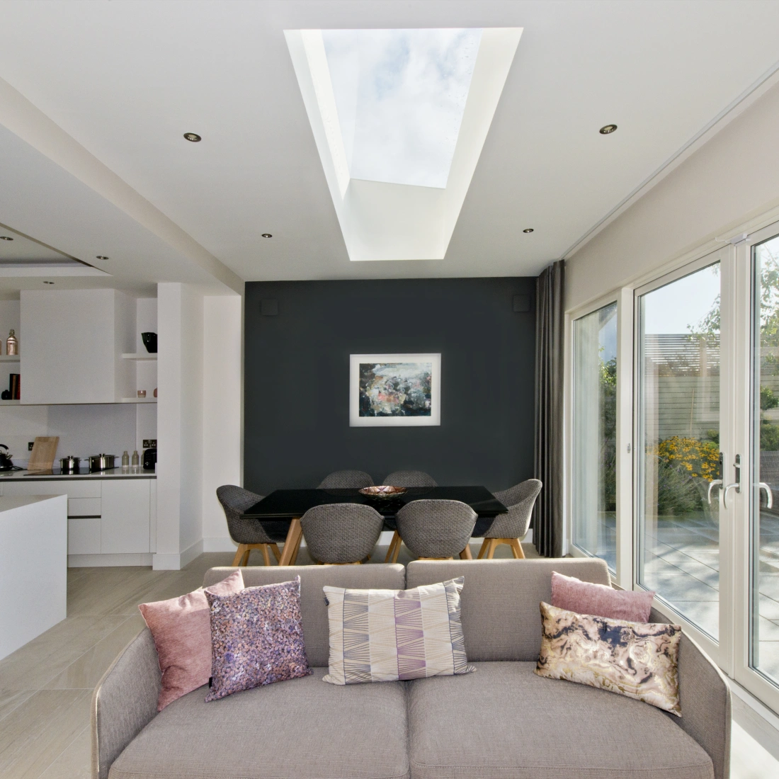 A modern interior showcasing an open living space with a couch adorned with decorative pillows. In the background, a dining area features a black table surrounded by gray chairs, and large glass doors lead to a garden. A skylight above provides natural light, illuminating the room.