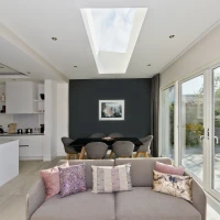 Un salon moderne avec un canapé beige orné de coussins colorés. À l’arrière, une salle à manger avec une table noire et des chaises grises. De grandes fenêtres et un plafond vitré laissent entrer la lumière naturelle, mettant en valeur les fleurs à l’extérieur.