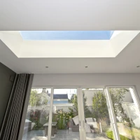 A modern room with a skylight that lets in natural light. The walls are painted a dark color, and large glass doors open to a garden with green plants and outdoor seating.