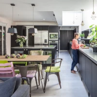 Cocina moderna con muebles oscuros y una mesa de comedor de madera. Una mujer de cabello recogido, vestida con una camisa de cuadros rojos y blancos, está de pie junto a la barra preparando alimentos. Hay plantas verdes y una ventana grande que aporta luz natural al espacio.