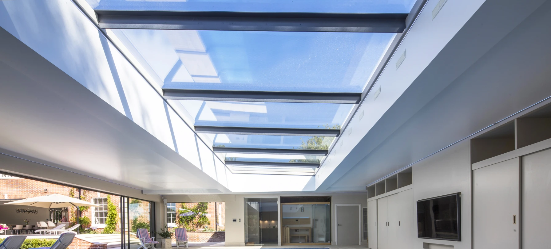A modern indoor swimming pool with a glass ceiling, featuring sleek white walls and a flat-screen TV mounted on the wall. Sunlight streams through the skylights, illuminating the water. Comfortable lounge chairs are arranged around the pool area.