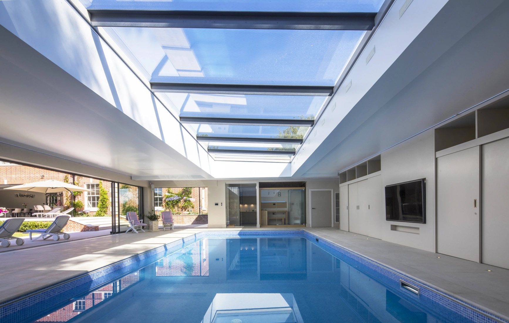 Innenansicht eines modernen, hellen Schwimmbades mit großen Fenstern und einem Glasdach. Das Wasser reflektiert das Licht. Es gibt Liegen am Rand des Beckens und einen Fernseher an der Wand. Die Umgebung wirkt elegant und einladend.