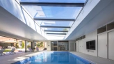 Innenansicht eines modernen, hellen Schwimmbades mit großen Fenstern und einem Glasdach. Das Wasser reflektiert das Licht. Es gibt Liegen am Rand des Beckens und einen Fernseher an der Wand. Die Umgebung wirkt elegant und einladend.