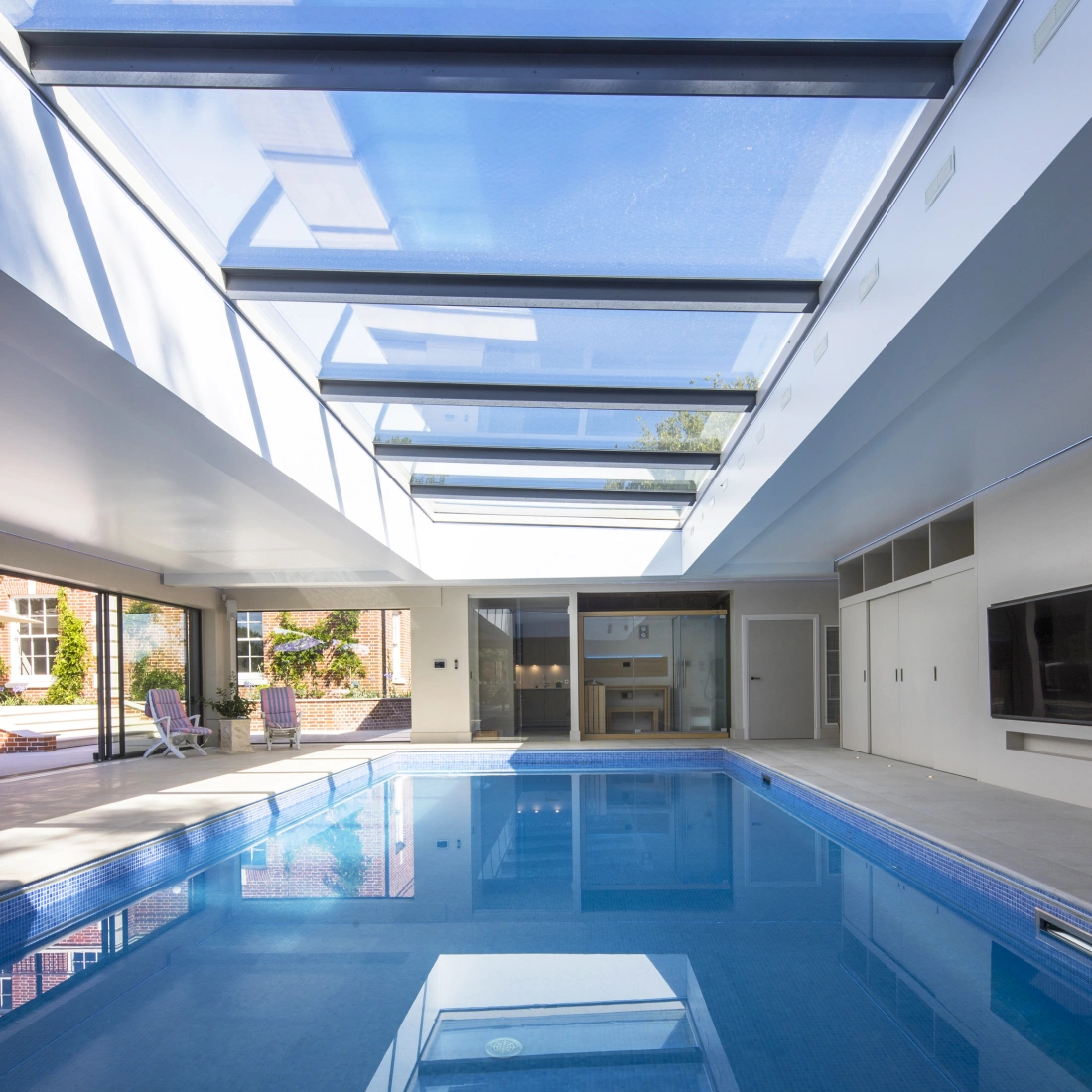 Interior de una casa con piscina iluminada por un techo de vidrio. Las paredes son de color claro y hay un área de descanso con sillas. Se puede ver el exterior con un jardín al fondo.
