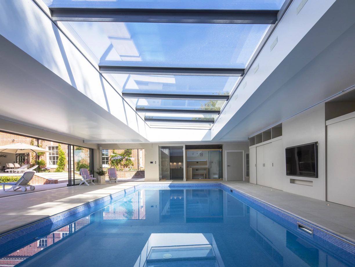 A modern indoor swimming pool with a glass ceiling, featuring sleek white walls and a flat-screen TV mounted on the wall. Sunlight streams through the skylights, illuminating the water. Comfortable lounge chairs are arranged around the pool area.