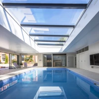 Intérieur d'une piscine moderne avec un plafond vitré, baignée de lumière naturelle. Des chaises longues sont disposées sur le côté et des portes coulissantes mènent à l'extérieur.