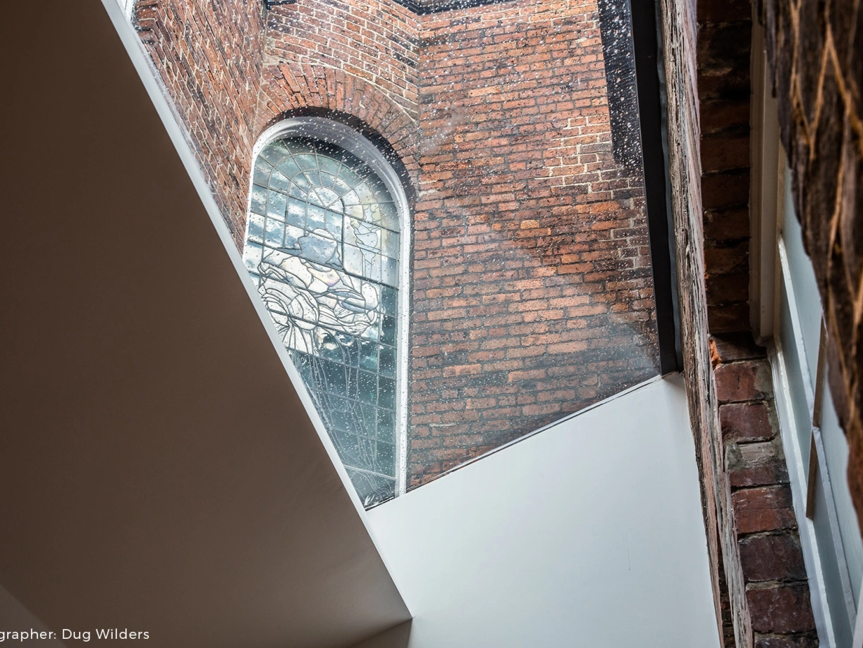Light streams through a large stained glass window, illuminating the textured red brick walls of an interior space. A modern, angled white surface contrasts with the historical architecture.