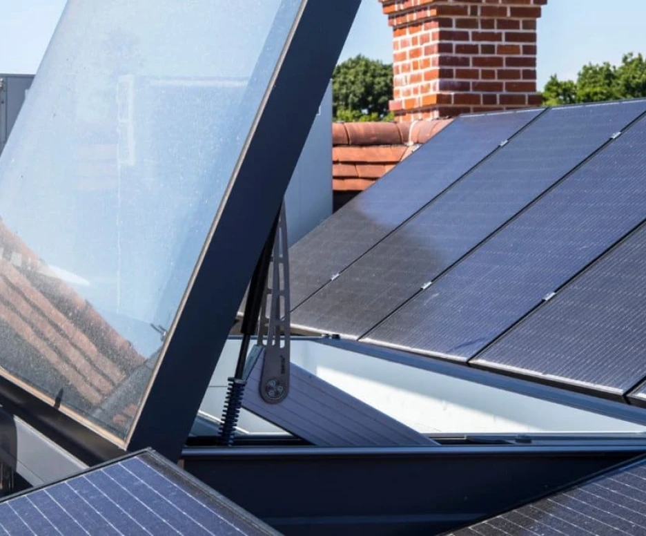 Solar panels installed on a rooftop, with one panel tilted open to expose a skylight or vent below. A red brick chimney is visible in the background, along with an air conditioning unit.