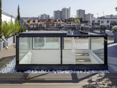 Toit-terrasse moderne avec une structure en verre et des plantes, offrant une vue sur la ville. Des galets blancs entourent la structure, et des immeubles se dressent en arrière-plan sous un ciel clair.