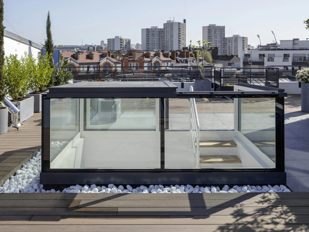 Toit-terrasse moderne avec une structure en verre et des plantes, offrant une vue sur la ville. Des galets blancs entourent la structure, et des immeubles se dressent en arrière-plan sous un ciel clair.