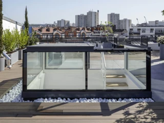 A modern rooftop terrace with a glass structure, surrounded by greenery and decorative white stones. In the background, city buildings and blue sky are visible.