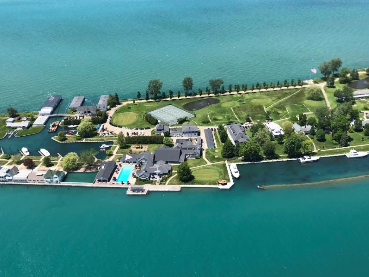 Aerial view of a lush, green island with waterfront properties, boats docked along the shore, and a swimming pool visible. The vibrant turquoise water surrounds the island, which features trees and neatly landscaped lawns.