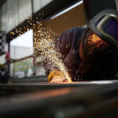 A person wearing a welding helmet and protective gear is working on metal, creating sparks as they weld. The background features industrial equipment and tools.
