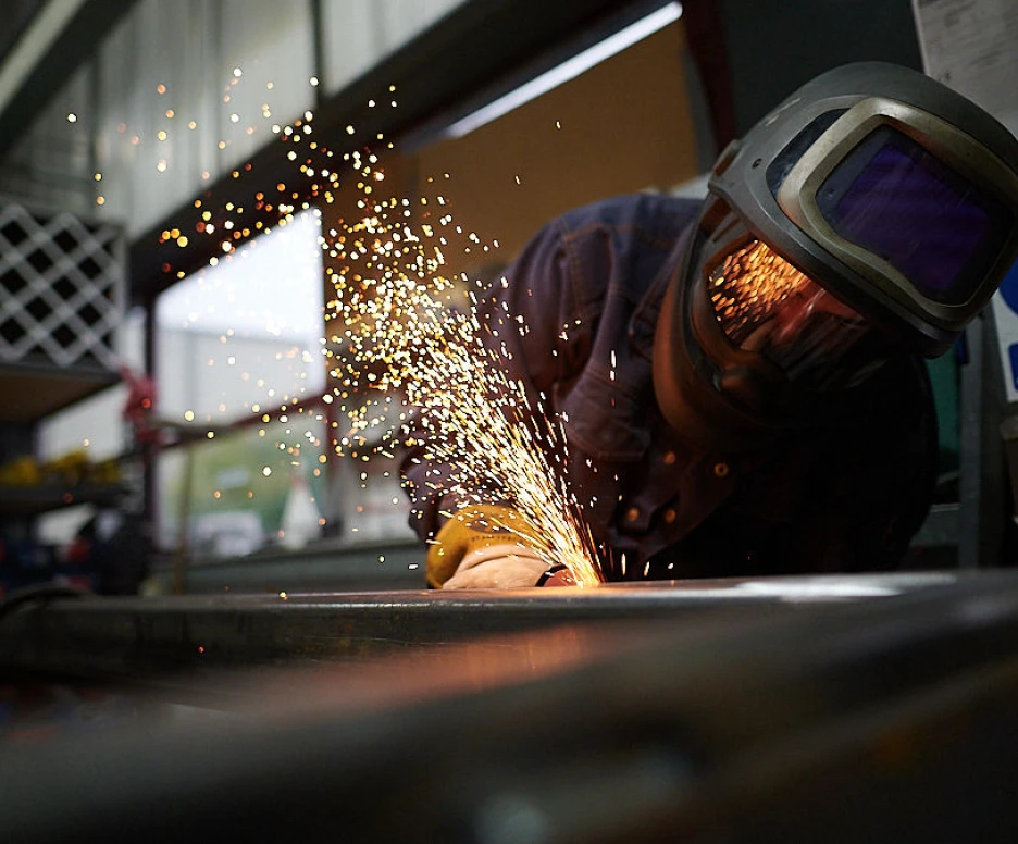 A welder wearing a protective helmet and gloves is working on metal, with sparks flying as they weld. The background shows a workshop with tools and equipment.