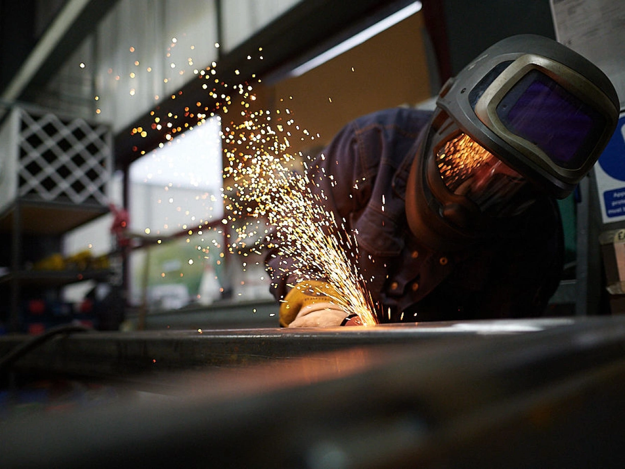 A welder wearing a protective helmet and gloves is working on metal, with sparks flying as they weld. The background shows a workshop with tools and equipment.