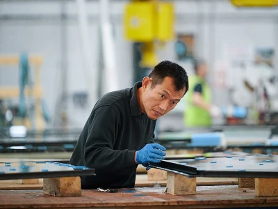 Un ouvrier travaille sur des panneaux de verre dans une usine. Il porte des gants bleus et se concentre sur sa tâche, tandis que d'autres personnes sont en arrière-plan.