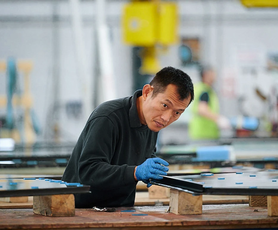 A worker in a factory setting inspects a large piece of glass. He is wearing blue gloves and focused on his task, with other workers and equipment visible in the background.