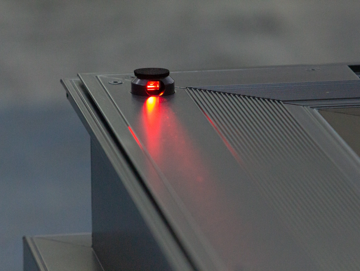 A close-up of a dark surface featuring a small, illuminated red button. The button is slightly raised with a textured surrounding.