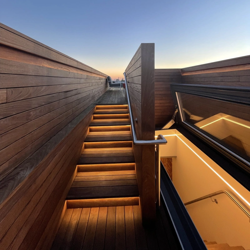 Wooden staircase with warm lighting leads upward, surrounded by wooden railings, against a clear evening sky transitioning to twilight. A glimpse of a modern structure is visible at the bottom of the stairs.
