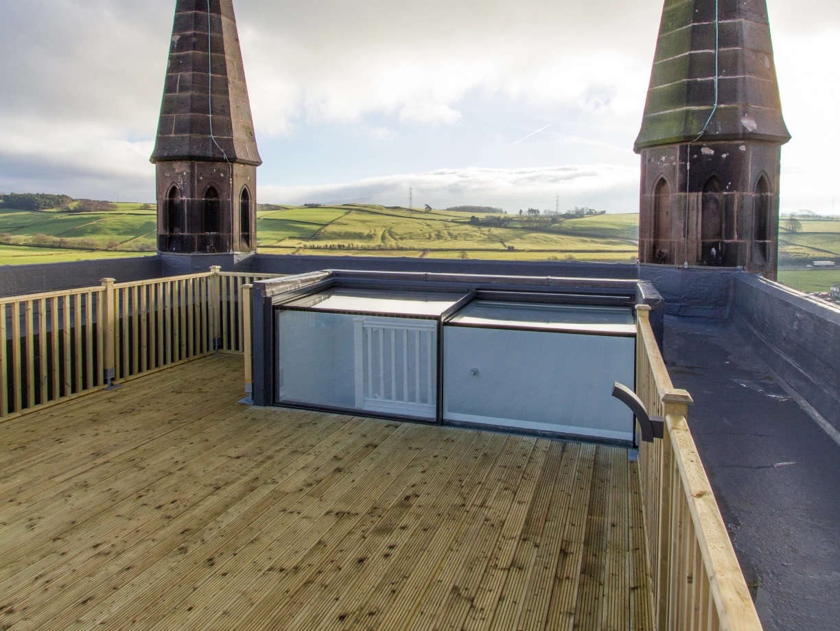A wooden rooftop deck offering a panoramic view of green hills, featuring two tall, pointed towers with a cloudy sky above.
