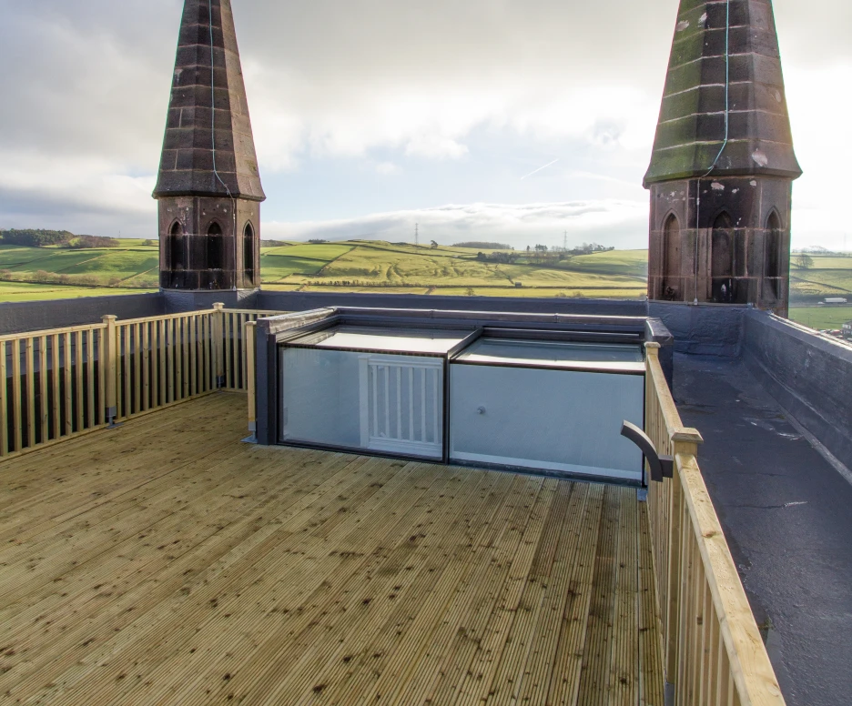 A rooftop view featuring two tall, pointed spires against a cloudy sky. Below, there's a wooden deck and a glass structure, with rolling green hills in the distance.