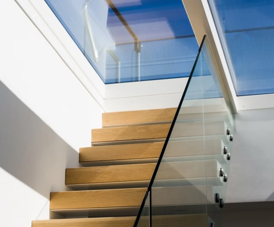 A modern staircase with wooden steps and a glass railing, illuminated by natural light streaming through a skylight above.