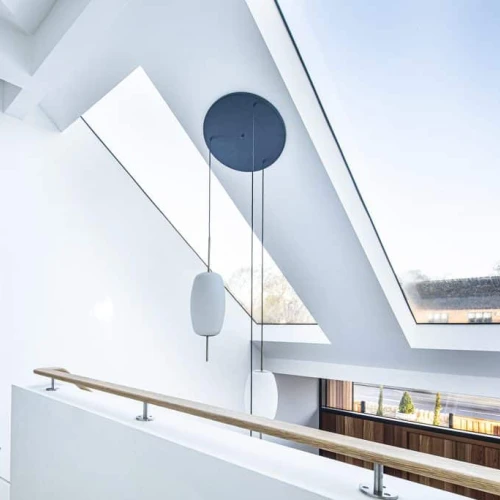 Heller Innenraum mit einer modernen Treppe und einem großen Dachfenster. Die Wände sind weiß, und es gibt eine Holzbalustrade. Ein kreisförmiger Pendelleuchter hängt an der Decke.