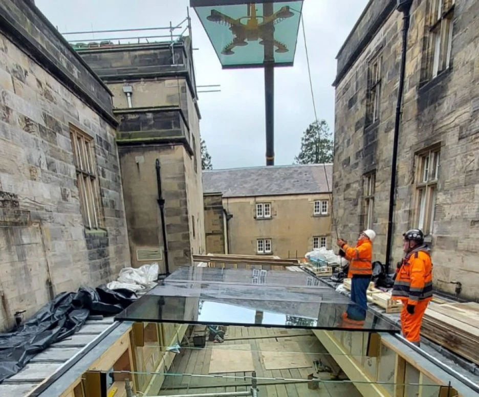Construction workers in safety gear stand on scaffolding around a newly constructed glass canopy, with an elaborate sign featuring an eagle in the background. The setting features stone buildings under a cloudy sky.