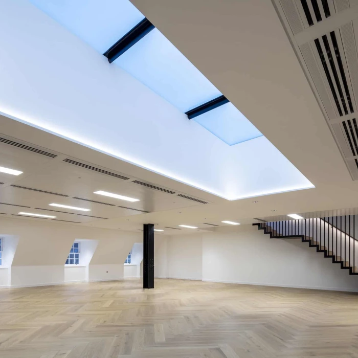 A spacious, modern room with wooden herringbone flooring. Features include a large skylight, recessed lighting, and a staircase along one wall. The walls are white, creating a bright and airy atmosphere.