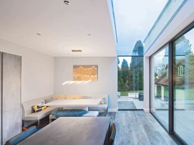 A modern dining area with a minimalist design features a built-in seating nook, a sleek table, and large glass windows showcasing a green outdoor space. Neutral colors dominate the interior, and a stylish light fixture hangs above the table.