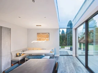 A modern dining area with a minimalist design features a built-in seating nook, a sleek table, and large glass windows showcasing a green outdoor space. Neutral colors dominate the interior, and a stylish light fixture hangs above the table.